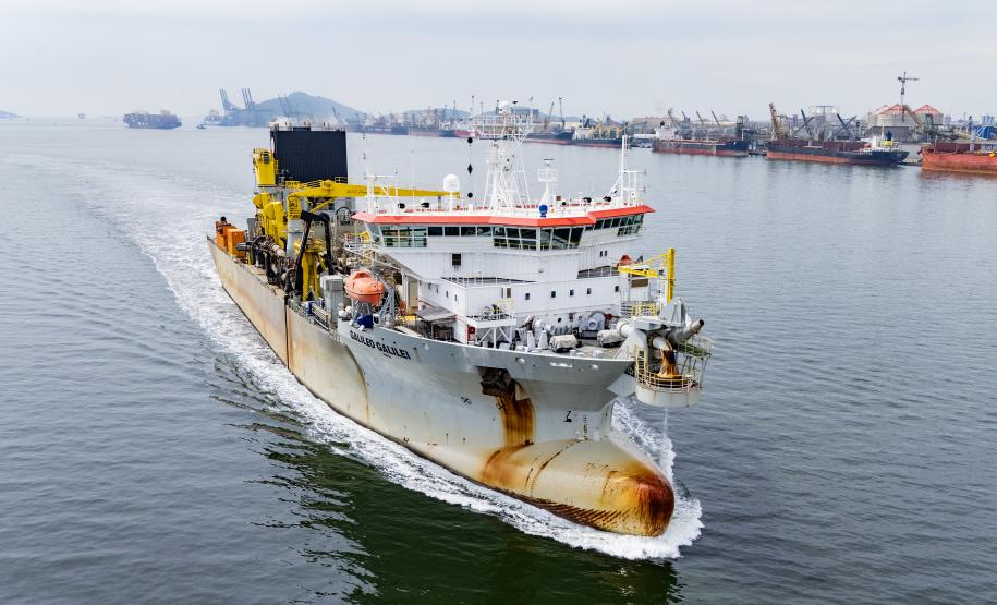 #ParaQuemNaoPodeVer Foto de um navio grande navegando em um corpo d'água, provavelmente próximo a um porto. O navio está indo em direção à direita da imagem e deixando um rastro na água. O navio é branco com detalhes amarelos e laranja, e parece equipado para operações industriais ou de transporte pesado, com guindastes e equipamentos grandes na parte traseira. Ao fundo, vê-se uma área portuária com guindastes, navios atracados e estruturas industriais próximas à água.