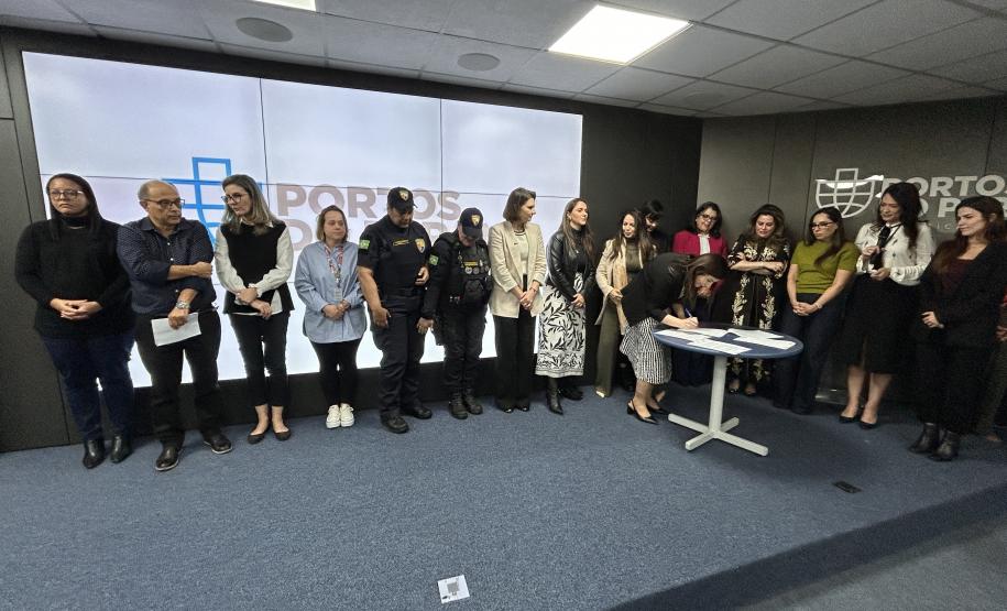 #ParaQuemNaoPodeVer Foto de um grupo de pessoas. São cerca de 19 pessoas, organizadas em duas fileiras, posando para uma foto em um ambiente interno, provavelmente uma sala de reuniões ou auditório. Ao fundo, há um painel com o logotipo e o nome “PORTOS DO PARANÁ”. Há uma mesa redonda branca e azul à frente do grupo. As pessoas estão vestidas de maneira variada, algumas de roupa social, outras de uniforme (aparentando ser de segurança).