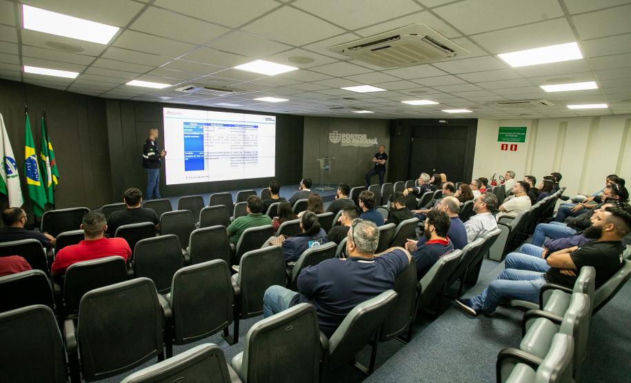 #ParaQuemNaoPodeVer Auditório com diversas pessoas sentadas em cadeiras assistindo a uma apresentação. Na frente, um homem está em pé próximo a um telão, onde uma apresentação de slides é projetada. Atrás dele, há bandeiras do Brasil e de outro estado ou instituição. O ambiente tem iluminação de teto forte, ar-condicionado e paredes claras. As pessoas parecem atentas à apresentação. A tela mostra informações em português, porém está levemente desfocada na imagem.