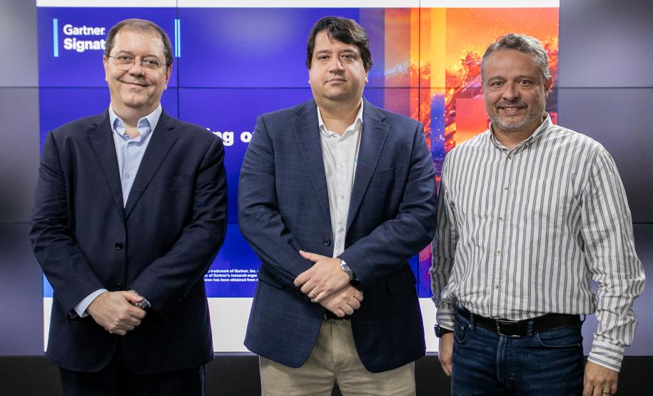 #ParaQuemNaoPodeVer Foto de três homens posando para a câmera. Eles estão de pé lado a lado. Dois deles usam ternos com camisa clara, e um, na frente, está sem paletó, usando uma camisa social listrada clara e jeans. Eles sorriem levemente. Ao fundo, aparece um painel iluminado com cores azuis e laranja, com parte da palavra “Gartner” visível.