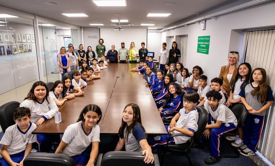 #ParaQuemNaoPodeVer Foto mostrando um grupo grande de pessoas sentadas ao redor de uma mesa de reunião em uma sala fechada. A maioria aparenta ser crianças e adolescentes, algumas usando uniformes escolares de calça azul e camiseta branca, enquanto adultos estão espalhados entre eles, talvez professores ou responsáveis. Todos olham para a câmera e sorriem, parecendo participarem de algum evento escolar ou visita guiada. No fundo, há quadros na parede e uma porta de vidro; do lado direito, há avisos e um car