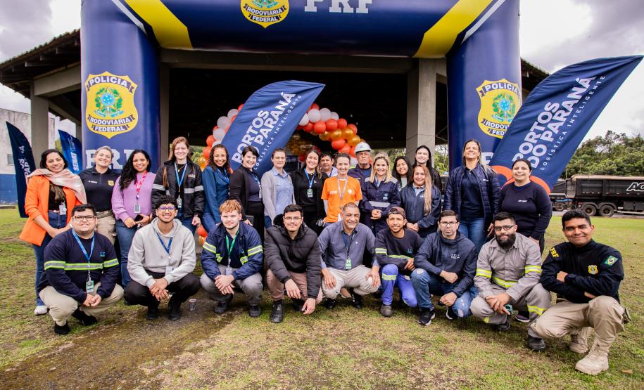 #ParaQuemNaoPodeVer Foto de um grupo grande de pessoas posando para a câmera, todas sorrindo, em frente a banners e bandeiras com os logotipos da Polícia Rodoviária Federal (PRF) e “Portos do Paraná – Logística. Eficiência. Valor.” Ao fundo, há balões laranja reunidos decorando a entrada de um prédio. As pessoas estão ao ar livre sobre uma área gramada, usam roupas variadas, incluindo uniformes da PRF, coletes e agasalhos. O ambiente é de evento institucional ou comemorativo, possivelmente uma ação conjunta