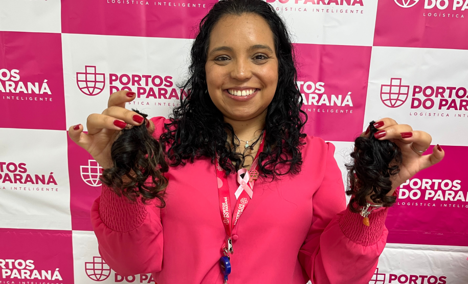 #praquemnaopodever foto mostra mulher negra de camisa rosa segurando uma mecha de cabelo de cada lado e sorrindo em frente de uma parede com os dizeres Portos do Paraná em rosa.