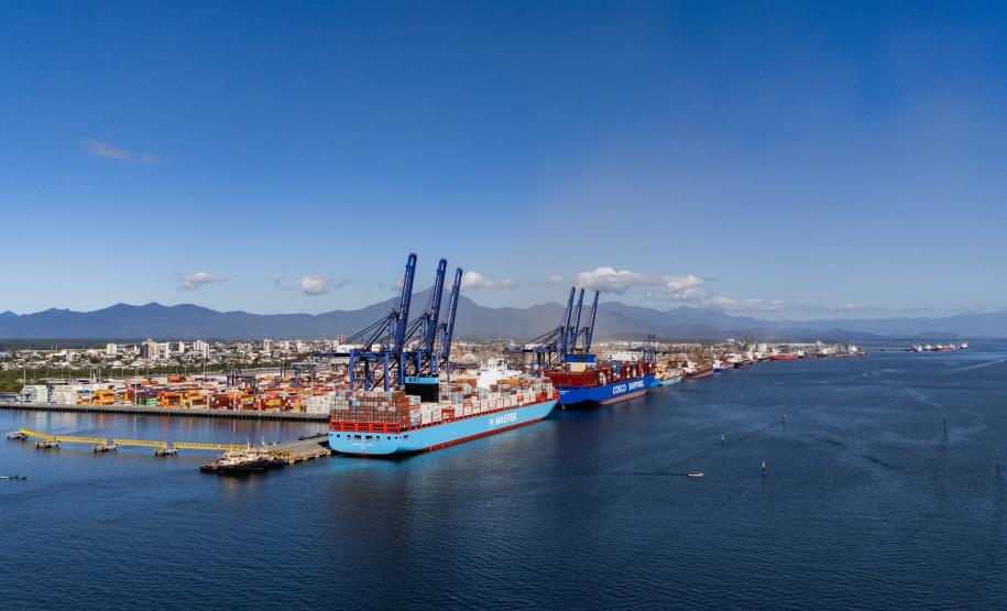 #ParaQuemNaoPodeVer Foto aérea de um porto. Na imagem, há um navio cargueiro grande, de cor azul, atracado em um píer. Próximo ao navio, vê-se diversos contêineres coloridos e guindastes altos, típicos de portos de carga. O mar ao redor é azul e há uma cidade ao fundo, com vários prédios e montanhas ao longe. A imagem está levemente inclinada para a direita.