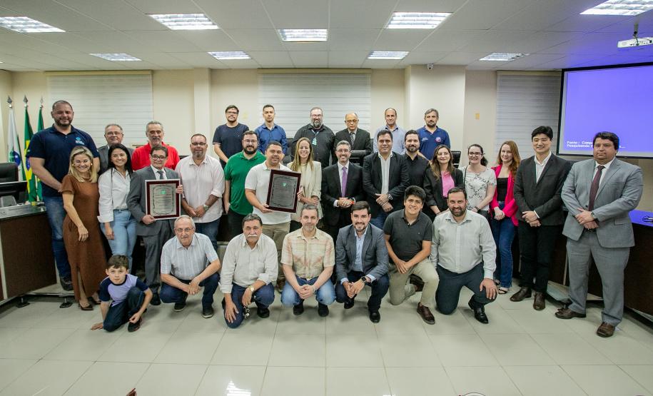 #ParaQuemNaoPodeVer Foto de um grupo grande de pessoas em um ambiente fechado, possivelmente uma sala de reuniões ou auditório. Todos estão vestidos de maneira formal e informal, sugerindo um evento corporativo ou de premiação. Algumas pessoas seguram placas ou certificados, indicando que receberam algum tipo de reconhecimento. Ao fundo, há bandeiras, reforçando o clima institucional do local. As pessoas estão sorrindo e posando para a foto, aparentando clima de celebração ou comemoração.