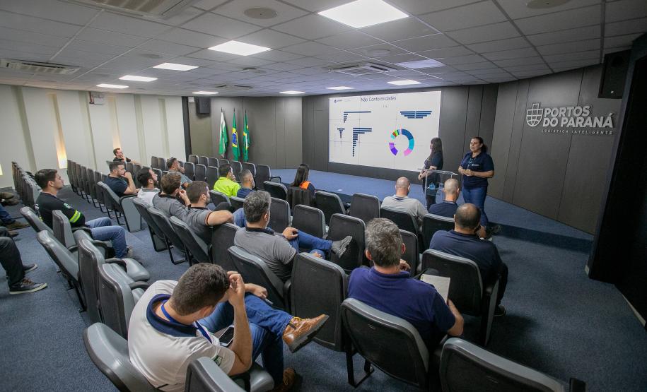 #ParaQuemNaoPodeVer Sala de apresentação com várias pessoas sentadas em cadeiras, assistindo a uma palestra. Na frente, há um telão exibindo gráficos e textos, além de duas pessoas apresentando. No fundo, há bandeiras do Brasil e do Paraná, e na parede está escrito "Portos do Paraná". O ambiente é bem iluminado, com teto branco e piso azul. A atenção das pessoas está voltada para o telão e os palestrantes.
