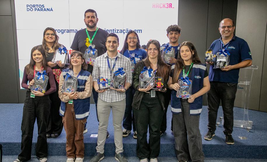 #ParaQuemNaoPodeVer Foto de um grupo de dez pessoas em pé, posando para a câmera em um ambiente interno, possivelmente em um evento ou cerimônia. Todas parecem estar sorrindo. Algumas seguram troféus e brindes, sugerindo premiação ou reconhecimento. O fundo tem um painel branco com texto, onde é possível ler “PORTOS DO PARANÁ”, “programa”, “digitalização”, e “HACK3R”. A maioria das pessoas usa crachás e roupas casuais ou uniformes.