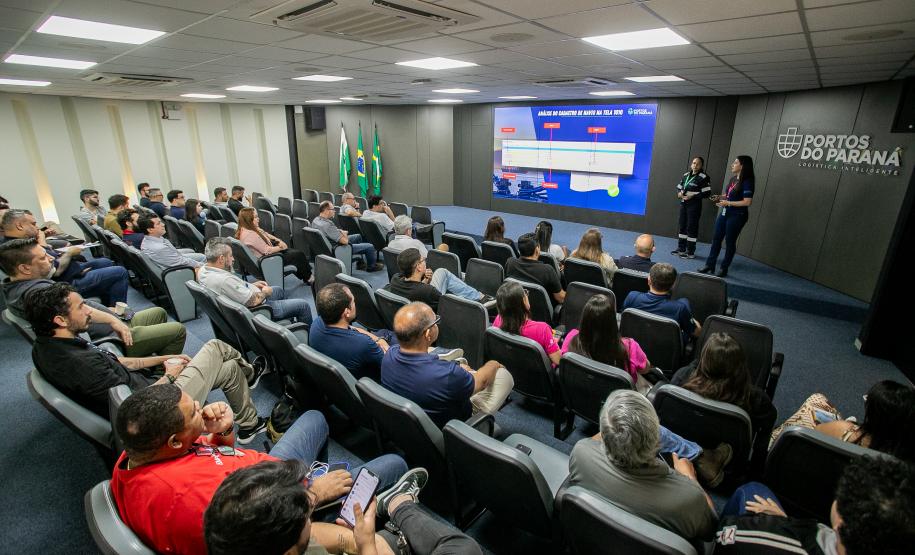 #ParaQuemNaoPodeVer Imagem de uma sala de conferências. Na foto, várias pessoas estão sentadas em poltronas, formando fileiras, e assistindo a uma apresentação feita por duas pessoas na frente da sala, próximo a um telão. No telão, há um slide azul com informações (textos e gráficos) e, na parede, o logotipo “Portos do Paraná” está visível. Também estão presentes bandeiras do Brasil.