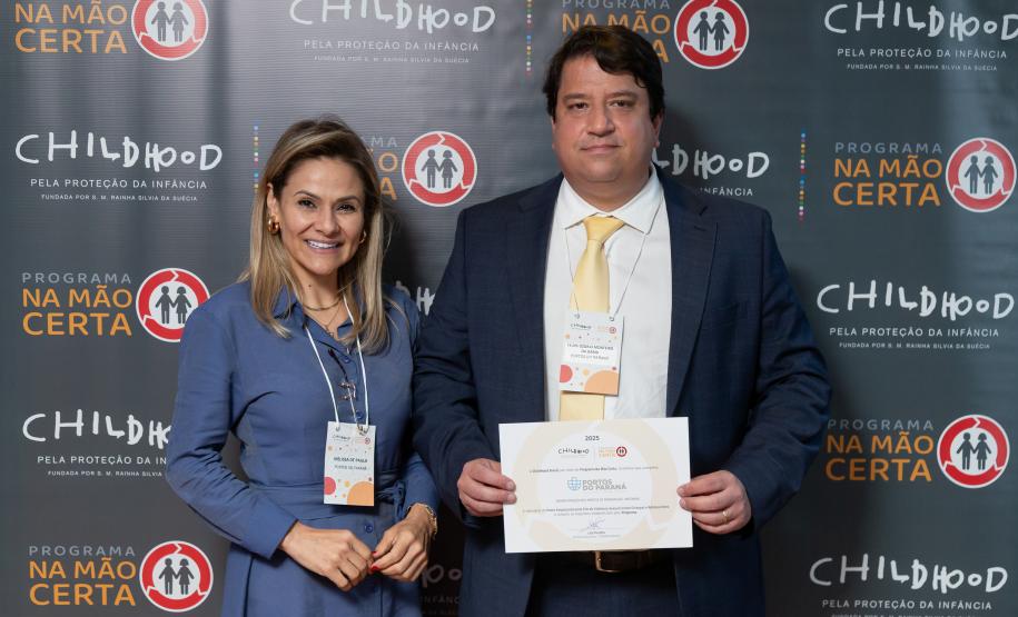 #ParaQuemNaoPodeVer Foto de um homem e uma mulher sorrindo, posando juntos para a câmera. Eles estão em frente a um painel de fundo decorado com os logos do "Programa Na Mão Certa" e da "Childhood Brasil", ambos destinados à proteção da infância. O homem está com um paletó azul, camisa branca e gravata amarela, segurando um certificado ou diploma. A mulher está usando uma roupa social azul claro, com crachá pendurado no pescoço. Ambos parecem estar participando de um evento formal ou solenidade de premiação