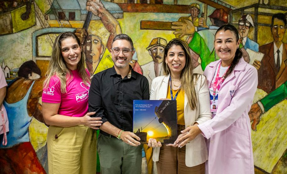 #praquemnaopodever foto mostra quatro pessoas lado a lado, em frente a um mural de trabalhadores, segurando um livro de capa azul e amarela escrito "Porto de Paranáguá - 90 anos"