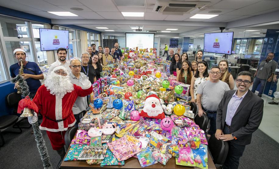 #praquemnaopodever a foto mostra uma mesa de madeira comprida e com quase mil brinquedos em cima de todas as cores e formatos. Nas lateriais, dezenas de pessoas estão a postos sorrindo e posando para o fotógrafo dentro de uma sala de reunião com tapete cinza e janelas grandes