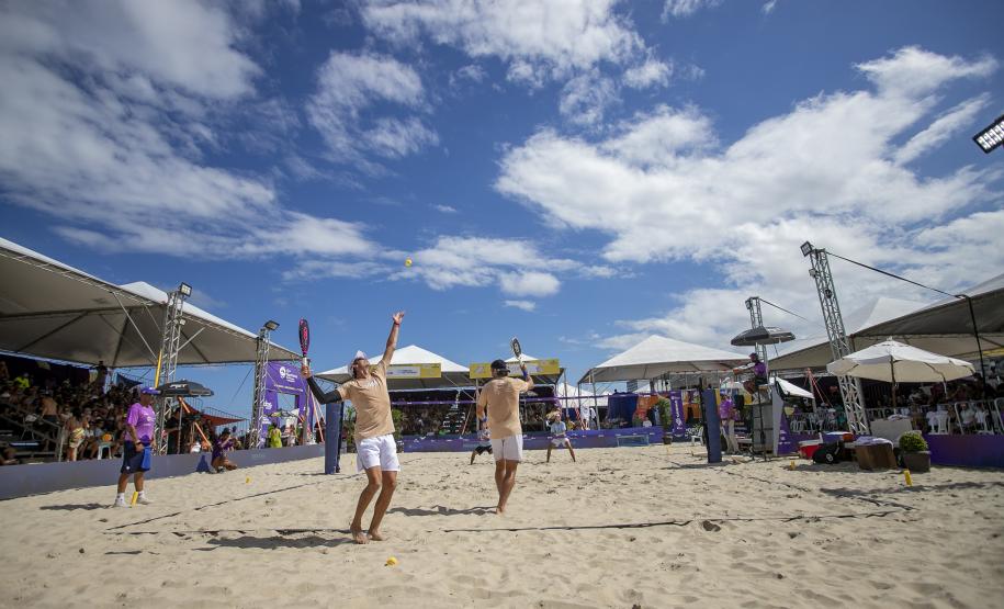 #praquemnaopodever imagem na praia, dentro da quadra de areia, a dupla de homens de bermuda branca e camisa amarela estão a frente de costas pra foto. Do outro lado está a outra dupla pronta pra receber a bola. O céu está azul com algumas nuvens. Ao fundo e dos lados as arquibancadas estão lotadas