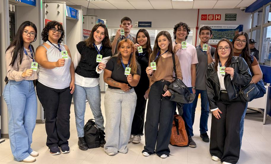 #ParaQuemNaoPodeVer Grupo de onze jovens alinhados em um corredor interno, todos segurando crachás verdes voltados para a frente. Na foto, as pessoas estão em pé, lado a lado, sorrindo. Há diversidade de tons de pele, de claros a médios e mais escuros, e estilos de cabelo diferentes, incluindo cabelos lisos, cacheados e ondulados, alguns longos, outros curtos, em cores como castanho, preto e loiro. As roupas são casuais: calças jeans claras e escuras, calça de moletom cinza, calça preta, tênis brancos