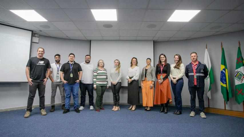 #ParaQuemNaoPodeVer Grupo de pessoas posando para uma foto em uma sala de reuniões. São 11 pessoas. Ao fundo, há três bandeiras: a do Brasil ao centro e duas verdes com detalhes vermelhos e um brasão, provavelmente de um estado ou município. O chão é acarpetado de azul e o teto tem placas brancas com iluminação embutida. As pessoas estão sorrindo e parecem estar em um evento formal ou institucional. 