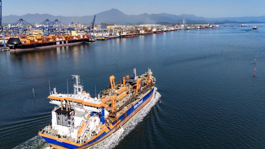 Uma draga navega pelo canal marítimo de acesso ao Porto de Paranaguá. A embarcação, nas cores branca, laranja e azul, aparece em primeiro plano deixando um rastro de espuma na água, indicando movimentação.  Ao fundo, é possível ver a estrutura do Porto de Paranaguá, com guindastes alinhados, contêineres empilhados e um navio cargueiro atracado. Mais atrás, surgem as montanhas da Serra do Mar sob uma iluminação suave, característica do início da manhã ou do fim da tarde.