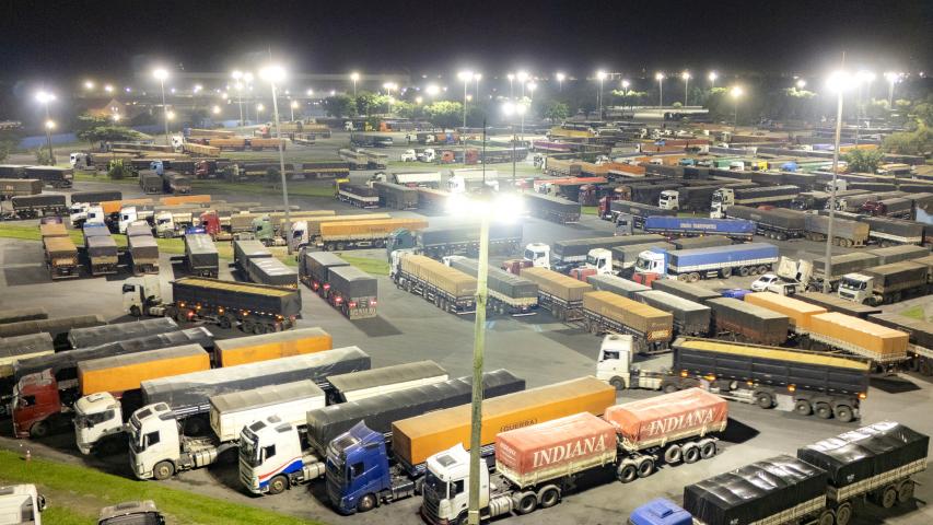 Imagem aérea noturna Pátio de Triagem de caminhões da Portos do Paraná. Vários caminhões e carretas estão estacionados lado a lado sob uma intensa luz que sai dos postes e super postes. 