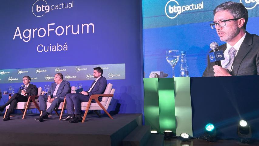 A imagem é de um palco onde há um grande telão azul ao fundo com o texto em letras grandes na vertical “AgroForum” e abaixo “Cuiabá”. Três homens de terno estão sentados em poltronas no palco, de frente para o público, com pequenas mesas de centro entre eles, onde há copos e garrafas de água.   No canto superior de outro telão está a imagem em destaque do diretor-presidente da Portos do Paraná.  Ele veste terno cinza e camisa branca e está segurando um microfone.  