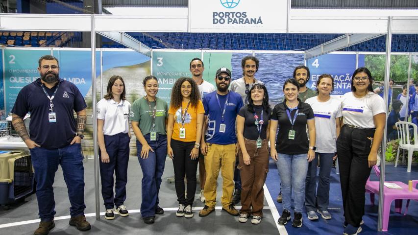 Há onze pessoas em pé, alinhadas em três fileiras, todas sorrindo para a câmera. Todos fazem parte da equipe de Meio Ambiente da Portos do Paraná. Eles estão posicionados na frente do stand da empresa.   