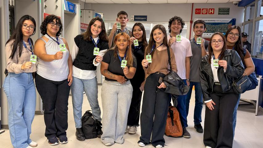 #ParaQuemNaoPodeVer Grupo de onze jovens alinhados em um corredor interno, todos segurando crachás verdes voltados para a frente. Na foto, as pessoas estão em pé, lado a lado, sorrindo. Há diversidade de tons de pele, de claros a médios e mais escuros, e estilos de cabelo diferentes, incluindo cabelos lisos, cacheados e ondulados, alguns longos, outros curtos, em cores como castanho, preto e loiro. As roupas são casuais: calças jeans claras e escuras, calça de moletom cinza, calça preta, tênis brancos